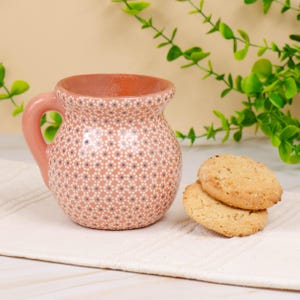 May include: A pink ceramic mug with a handle, adorned with a white floral pattern. Two golden-brown cookies rest beside the mug on a white surface. Green foliage is in the background.