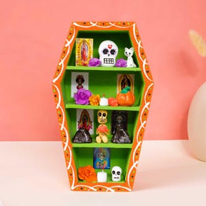 May include: A coffin-shaped wooden shelf painted orange with white decorative accents and a bright green interior. The four-tiered display holds various miniature figurines, including skulls, small religious cards, artificial flowers, and a cat.