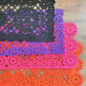 May include: Four layered paper doilies in black, purple, pink, and orange. The doilies have intricate cut-out designs of flowers and skulls.