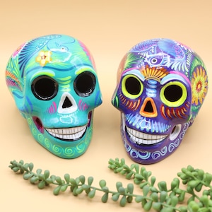 May include: Two colorful ceramic sugar skulls, one teal with pink and green floral designs and the other purple with yellow and orange floral designs.