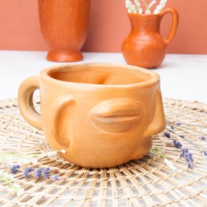May include: A terracotta-colored ceramic mug with a face design, featuring a nose and closed lips. The mug has a handle and sits on a woven mat, with other terracotta pottery in the background. Dried lavender sprigs add a decorative touch.