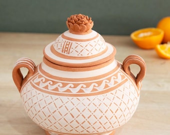 May include: A terracotta ceramic jar with a lid, featuring a floral knob. The jar is painted with a white and brown geometric pattern, including a wave design. It has two handles and is set on a wooden surface.