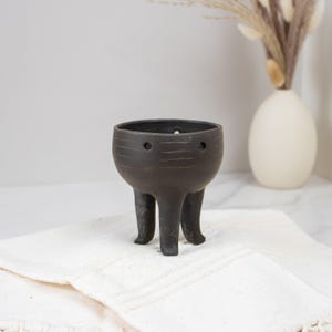 May include: A small, dark gray ceramic planter with three legs. The planter has a round body with horizontal etched lines and two small holes near the top. The planter is sitting on a white cloth.