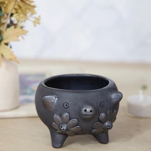 May include: A dark gray ceramic planter shaped like a pig, with a round body, small legs, and a detailed face. The planter features daisy-like flower accents and is designed for small plants or succulents.