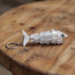 May include: A fish-shaped keychain crafted from iridescent white mother-of-pearl scales layered to form the body. The fish features a small dark eye, an open mouth, and a metal ring attached to the tail fin by a short silver-toned chain link.