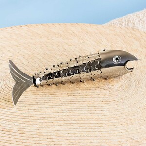 May include: A silver-toned metal fish-shaped bottle opener with a segmented, scale-like body design. The object features a smooth head with a circular eye detail, a wide tail fin, and a functional opening mechanism located at the mouth area.