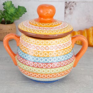 May include: A colorful ceramic sugar bowl with a lid. The bowl is decorated with stripes and dots in shades of orange, yellow, green, blue, and red. The lid has a knob on top.