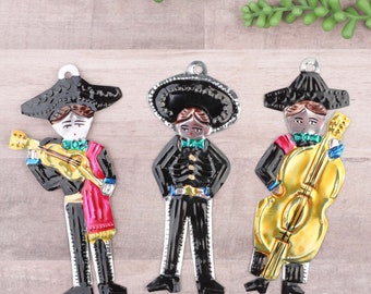 May include: Three metal ornaments depicting figures in traditional Mexican mariachi attire. The figures are playing a guitar, a trumpet, and a cello. The ornaments are painted in black, red, yellow, and white.