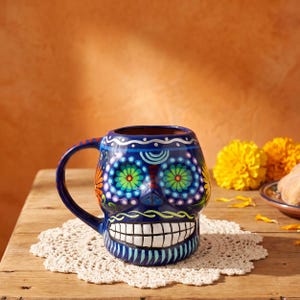 May include: A dark blue ceramic mug shaped like a skull, featuring vibrant hand-painted floral patterns in green, yellow, and orange around the eye sockets. The mug has a wide handle and a prominent white toothy grin painted across the lower section.