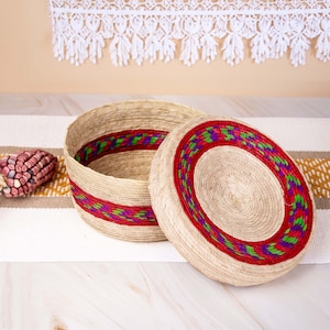 May include: A round, lidded storage basket made of natural tan woven fibers. The basket and its lid feature a decorative band of vibrant red, purple, and green woven patterns. The container has a flat base and a matching circular lid.