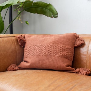 May include: A terracotta-colored throw pillow with a woven pattern and tassels on the corners. The pillow is on a brown leather couch.
