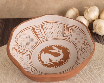 May include: A terracotta bowl with a scalloped rim featuring a white painted rabbit in the center. The interior walls are decorated with white geometric cross-hatch patterns and leaf motifs against the natural reddish-brown clay background.