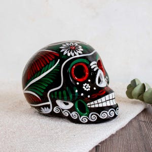 May include: A black ceramic skull figurine featuring hand-painted floral and swirl patterns in red, green, and white. The skull has prominent red eye sockets, a white nasal cavity, and a row of white teeth, with a large white flower on top.