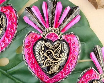 Könnte beinhalten: Dekorative herzförmige Zinnornamente in leuchtendem Pink und Schwarz. Jedes Ornament hat ein zentrales Herzdesign mit einem Vogel- und Blumenmotiv, akzentuiert mit goldenen Details. Die Ornamente haben einen strukturierten, geprägten Rand und eine fächerförmige Oberseite.