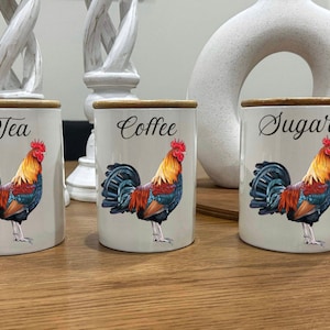 May include: A trio of ceramic kitchen canisters for tea, coffee, and sugar. Each white canister has a bamboo lid and a colourful rooster illustration. The words "Tea", "Coffee", and "Sugar" are written on the front.