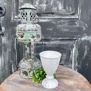 May include: A distressed green metal lantern with a glass insert and a white metal planter with a distressed finish. The planter is sitting on a wooden table with a small bouquet of white flowers.