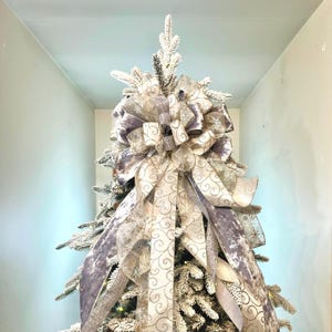 May include: A Christmas tree topper featuring a large bow made of silver and cream-colored ribbons. The ribbons have a swirl pattern and are accented with glitter. The tree is flocked with white, creating a wintery look.