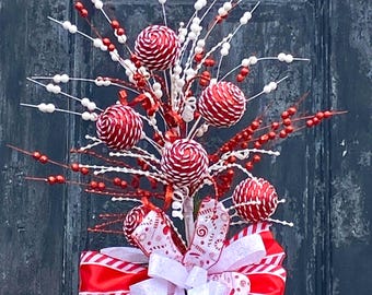 May include: A festive Christmas wreath with red and white striped ball ornaments, white berry sprigs, and a large red and white striped bow. The wreath is set against a dark blue door.