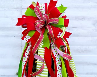May include: A festive Christmas tree topper featuring a large bow with red, green, and white ribbons. The bow has a mix of solid, polka dot, and striped patterns. The topper is designed to sit atop a Christmas tree.