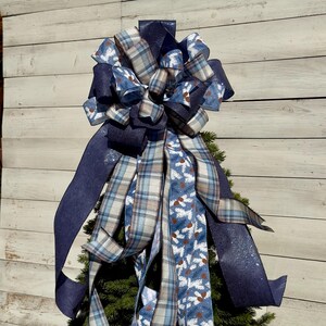 May include: A blue, white, and brown plaid ribbon, a blue ribbon with a white snowflake pattern, and a dark blue ribbon with glitter are tied together in a large bow. The bow is on top of a green Christmas tree.