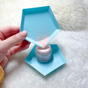 May include: A light blue plastic tray with pink powder being poured into a small clear jar with a white lid. The jar is sitting on a light blue plastic tray.