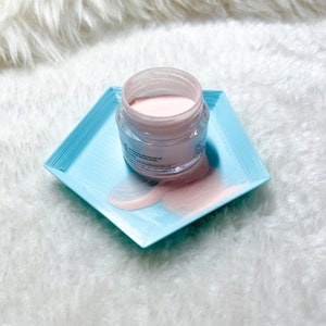 May include: A small, clear jar containing pink powder. The jar is on a light blue, square-shaped tray. The powder is spilling out of the jar onto the tray.