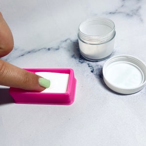 Könnte beinhalten: Ein rosa Plastikbehälter mit weißem Pulver darin. Ein Finger taucht in das Pulver. Im Hintergrund befindet sich ein durchsichtiges Plastikglas mit weißem Deckel.