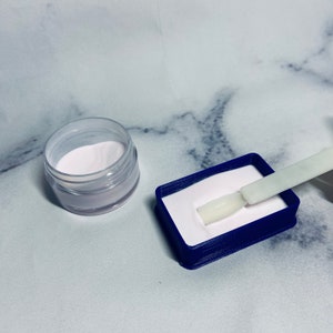 May include: A small clear jar filled with white powder and a blue plastic container with a white powder and a white plastic spatula.