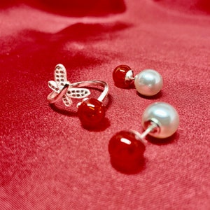 Könnte beinhalten: Ein silberner Ring mit Schmetterlingsdesign und zwei Paar Ohrringe. Die Ohrringe haben eine rote Perle gepaart mit einer weißen Perle. Der Schmuck wird auf einer roten Satin-Oberfläche präsentiert.