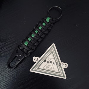 May include: Black and green paracord keychain with a black metal clip and a silver ring. The keychain has a sticker with the text "VET PARACORD MMXX".