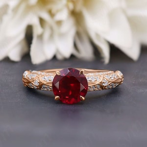 May include: A rose gold engagement ring with a large, round, red ruby gemstone surrounded by smaller, round, white diamonds. The band is decorated with a delicate, floral design.
