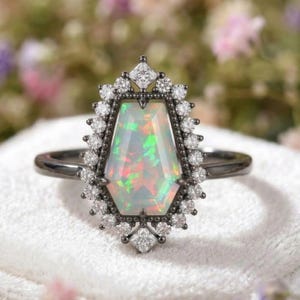 May include: A black metal ring featuring a large, iridescent opal gemstone in a unique geometric shape. The opal is surrounded by a halo of small, sparkling diamonds. The ring's band is a simple, dark metal.