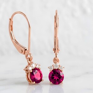 May include: Rose gold leverback earrings with oval, deep red gemstones. Each earring features a cluster of small, clear stones above the main gem. The earrings are displayed against a soft, white background.