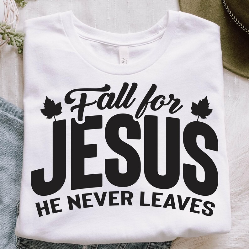 May include: White t-shirt with the text "Fall for JESUS He Never Leaves" in black. The design includes leaf accents. The shirt is folded and displayed on a white surface.