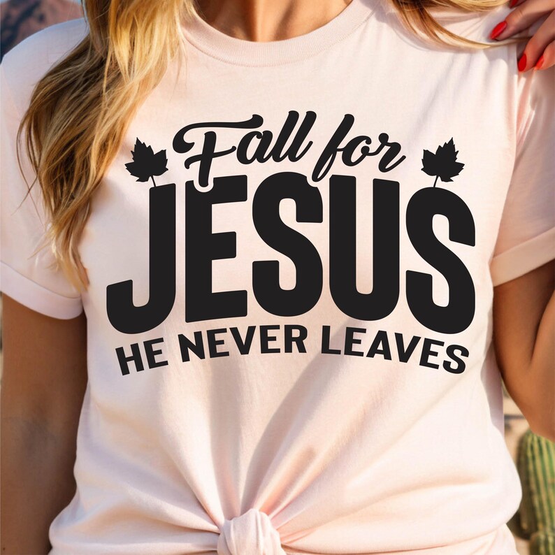 May include: A light pink t-shirt with the text "Fall for JESUS HE NEVER LEAVES" in black. The text is stylized with decorative leaf accents. The shirt has short sleeves and a knotted front.