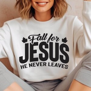May include: Cream-colored sweatshirt with the text "Fall for JESUS, HE NEVER LEAVES" in black. The text has a decorative font with maple leaf accents. The sweatshirt is a casual style.