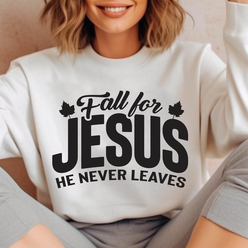 May include: Cream-colored sweatshirt with the text "Fall for JESUS, HE NEVER LEAVES" in black. The text has a decorative font with maple leaf accents. The sweatshirt is a casual style.