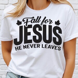 May include: White t-shirt with the text "Fall for JESUS" in a black, stylized font, with leaf accents. Below, the text "HE NEVER LEAVES" is also in black. The shirt is a casual, short-sleeved style.