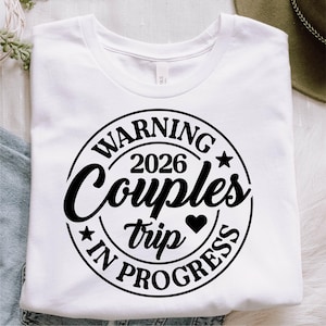 May include: White t-shirt with a black circular graphic. The graphic reads "WARNING 2026 Couples trip IN PROGRESS" with stars and a heart. The shirt is folded and appears to be made of a soft material.