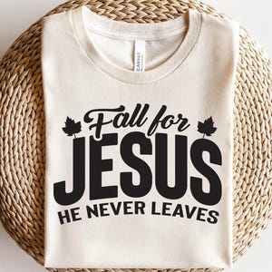 May include: Cream-colored t-shirt with the text "Fall for JESUS, He Never Leaves" in black. The design includes leaf accents. The shirt is made of a soft material and is displayed on a woven surface.