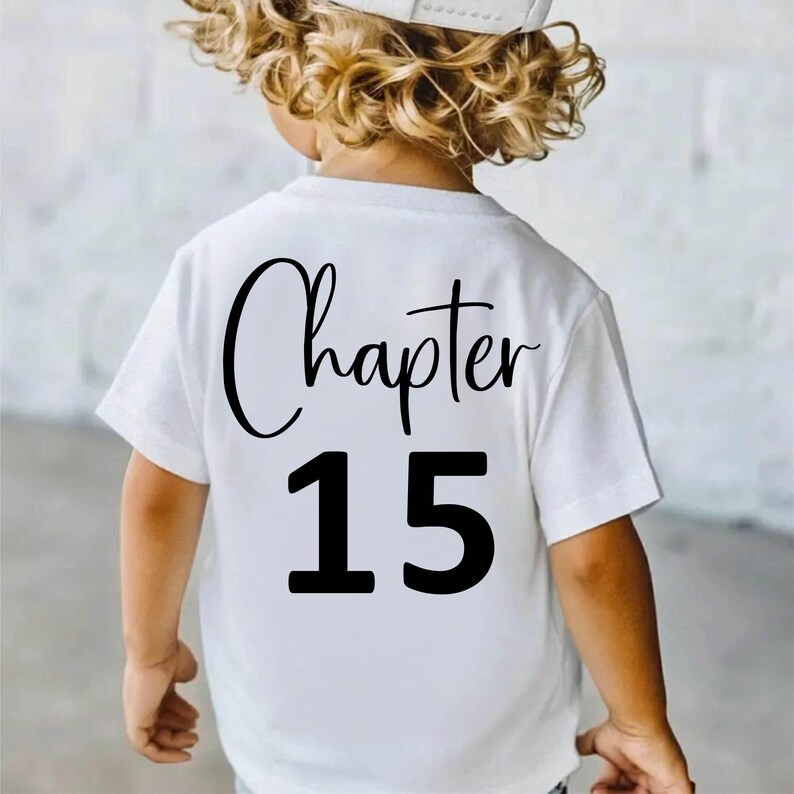 May include: White t-shirt with the words "Chapter 15" printed in black. The word "Chapter" is in a cursive font, and the number "15" is in a bold, block font. The shirt is worn by a child.