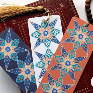 May include: Three colourful bookmarks with intricate geometric designs featuring blue, teal, orange, and gold accents. Each bookmark has a gold tassel.