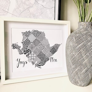 May include: A black and white framed print of a map of an island with a geometric pattern. The island is labeled "Ynys Môn" in black text.