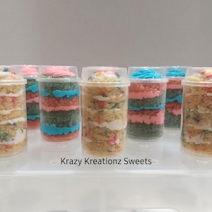 May include: Six clear plastic containers with lids, each containing a layered cake pop. The cake pops are decorated with sprinkles and frosting in pink, blue, and green. The containers are arranged in a row on a white surface. The text "Krazy Kreationz Sweets" is visible below the containers.