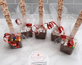 May include: Five square chocolate blocks on wooden sticks, each wrapped in clear plastic with red ribbon. The sticks feature a repeating brown pattern of mugs labeled cocoa and marshmallows. Toppings include colorful candies and white marshmallows.