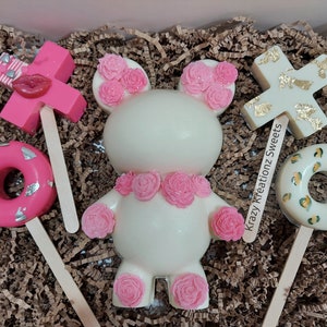 Może przedstawiać: Pudełko z dekoracyjnymi słodyczami, w tym biała słodycz w kształcie misia z różowymi akcentami róż, oraz różowe i białe smakołyki na patyczkach. Smakołyki obejmują "X" ze złotymi liśćmi, pączka i "X" z motywem ust.