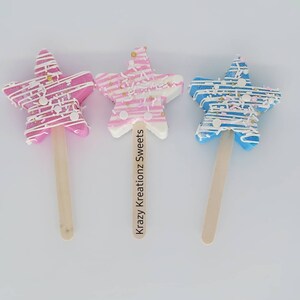 Könnte beinhalten: Drei sternförmige Cake Pops auf Holzstielen. Die Cake Pops sind mit weißen und rosa Streifen, weißen und blauen Streifen und weißen und blauen Streifen mit goldenen Streuseln verziert. Die Cake Pops befinden sich auf einem weißen Hintergrund. "Krazy Kreationz Sweets" steht auf dem Stiel des mittleren Cake Pops.
