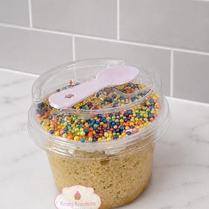 May include: A clear plastic container filled with a light-colored cake, topped with a layer of multi-colored sprinkles and a light purple plastic spoon. A white label on the side features a cupcake icon and the text Krazy Kreationz Sweets.