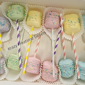 Puede incluir: Una caja de paletas de malvavisco de colores pastel decoradas con chispas de colores. Las paletas están en palos y están dispuestas en una caja con forros de papel blanco. La caja tiene el texto "Krazy Kreationz Sweets" impreso en ella.
