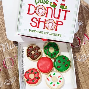 May include: A box of decorated cookies resembling donuts. The box is labeled "North Pole Donut Shop" with elf-themed graphics. The cookies are frosted in various colors, including red, green, brown, and white, with sprinkles and icing details. "Overnight Elf Delivery" is also printed on the box.